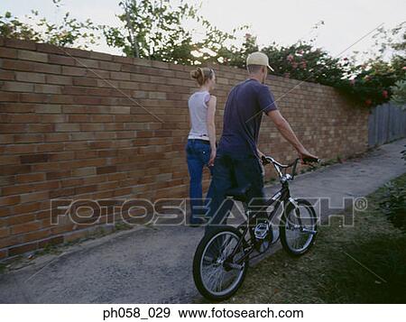 Stock Photo - Young man and woman walking with bike, rear view. Fotosearch