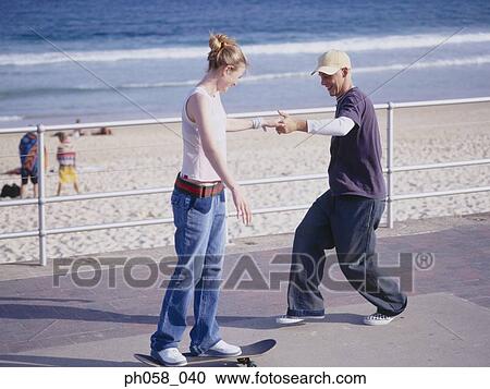 Stock Image - Young man helping young woman balancing on skateboard. Fotosearch
