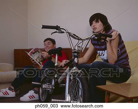 Stock Image - Couple sitting on sofa, young woman supporting bike. Fotosearch