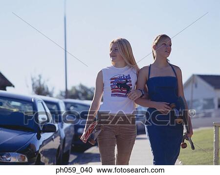 Stock Image - Two women walking on walkway holding skateboards . Fotosearch