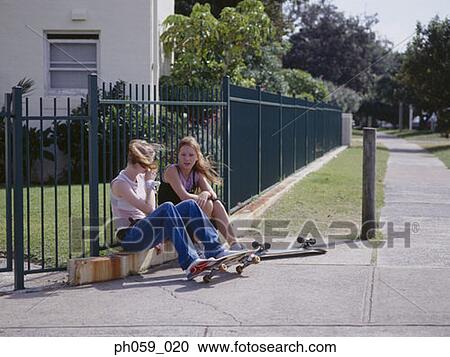 Stock Image - Two young women sitting on curb, chatting. Fotosearch