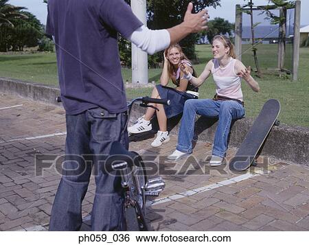 Stock Photograph - Young man holding bike talking to young women sitting on curb. Fotosearch