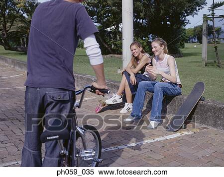 Young man holding bike talking to young women sitting on curb View Large Photo Image Stock Photography - Young man holding bike talking to young women sitting on curb. Fotosearch
