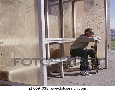 Stock Photo - Young man sitting at bus stand holding a skateboard. Fotosearch