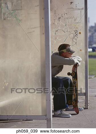 Stock Photo - Young man sitting at bus stand holding a skateboard. Fotosearch