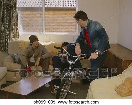 Stock Photography - Young man sitting on bike, while friends gather on sofa. Fotosearch