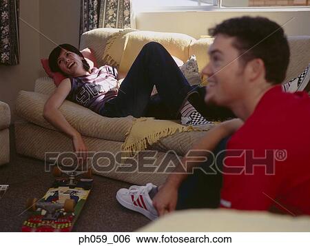 Stock Photograph - Young woman lying on sofa, while friend on sofa. Fotosearch