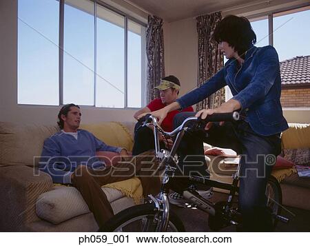 Stock Image - Young woman sitting on bike, while friends gather on sofa. Fotosearch