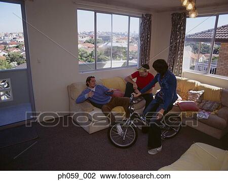 Stock Image - Young woman sitting on bike, while friends gather on sofa. Fotosearch