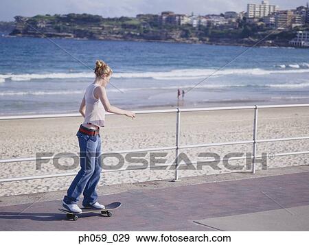 Stock Photo - Young woman skateboarding on pathway, near beach. Fotosearch