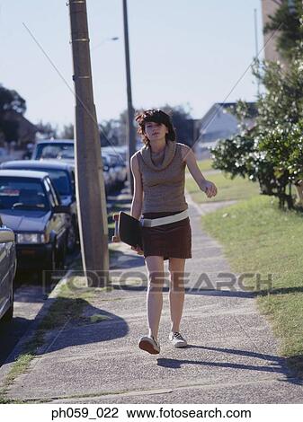 Stock Image - Young woman walking on walkway, holding a skateboard. Fotosearch