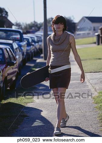 Stock Image - Young woman walking on walkway, holding a skateboard. Fotosearch