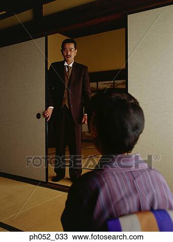 Stock Image - Japanese woman sitting in room, man standing by door. Fotosearch