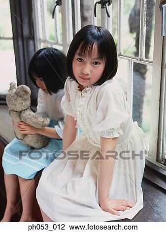 Stock Image - Two girls sitting on window ledge, portrait. Fotosearch