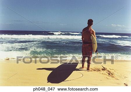 Young man on beach holding surfboard, rear view View Large Photo Image Stock Photo - Young man on beach holding surfboard, rear view. Fotosearch