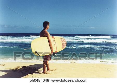 Young man on beach holding surfboard, rear view View Large Photo Image Stock Photo - Young man on beach holding surfboard, rear view. Fotosearch