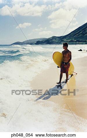 Young man walking along a beach with surfboard View Large Photo Image Picture - Young man walking along a beach with surfboard . Fotosearch
