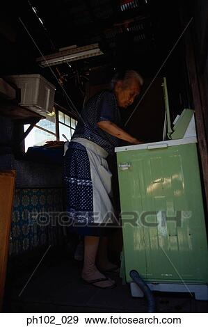 Stock Photo - Matured Japanese woman places laundry in washing machine. Fotosearch
