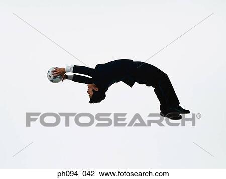 Stock Image - Young businessman leaping in air, holding a soccer ball. Fotosearch