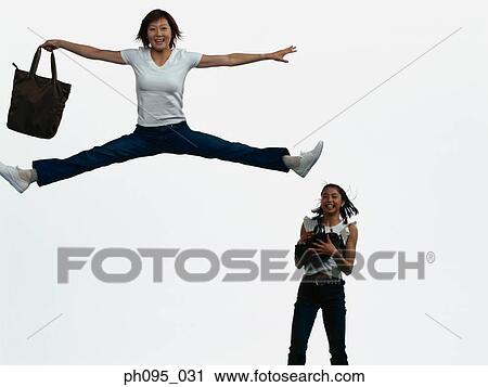 Stock Image - Two young Asian women in jeans & t-shirt, carrying bags, jumping in mid-air. Fotosearch
