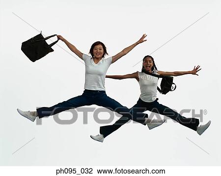 Stock Image - Two young Asian women in jeans & t-shirt, carrying bags, jumping in mid-air. Fotosearch