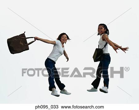 Stock Image - Two young Asian women in jeans & t-shirt, carrying bags, jumping in mid-air. Fotosearch