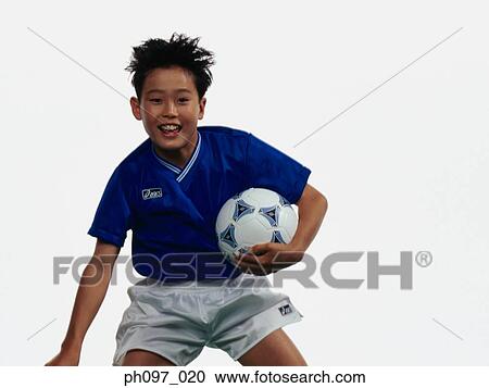 Stock Image - Young Asian boy holding football, jumping in mid-air. Fotosearch