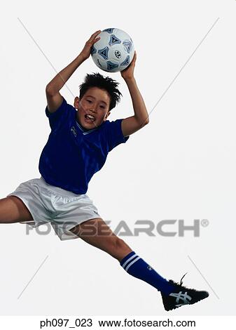 Stock Image - Young Asian boy holding football, jumping in mid-air. Fotosearch