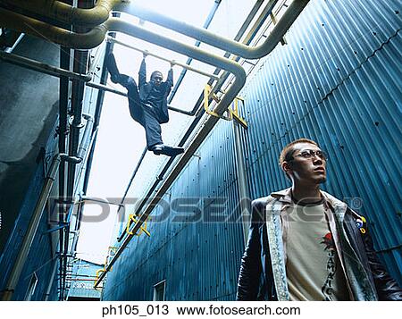 Stock Image - Man hanging on pipes, while man walking ahead. Fotosearch