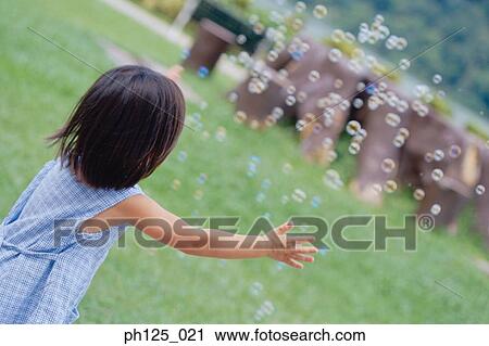 Stock Image - Young Asian girl playing with bubbles, rear view. Fotosearch