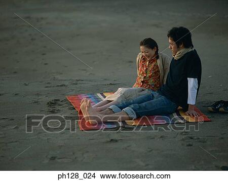 Picture - Young couple sitting on a rainbow mat on the beach. Fotosearch