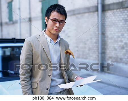 Asian Man with documents leaning on a car View Large Photo Image Picture - Asian Man with documents leaning on a car. Fotosearch