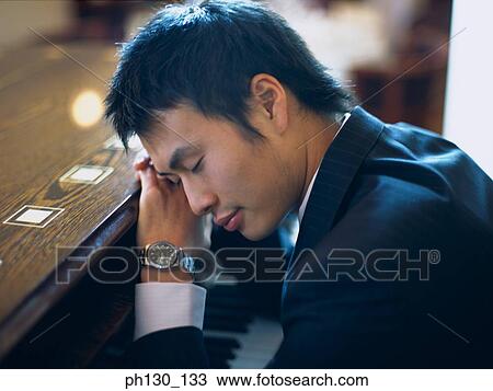 Stock Image - Asian Man resting on a piano. Fotosearch