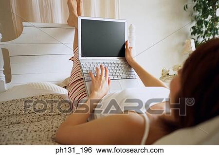 Stock Photograph - Woman lying on a bed with a laptop. Fotosearch