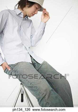 Young man sitting on a stool holding rim of his hat View Large Photo Image Stock Photograph - Young man sitting on a stool holding rim of his hat. Fotosearch
