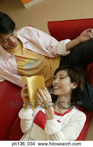Picture - Young couple lying on a couch reading a book. Fotosearch