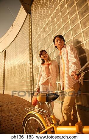 Stock Image - Young couple standing against a wall holding a bicycle. Fotosearch