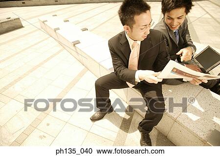 Stock Image - High angle view of two young businessmen sitting outdoors and talking. Fotosearch