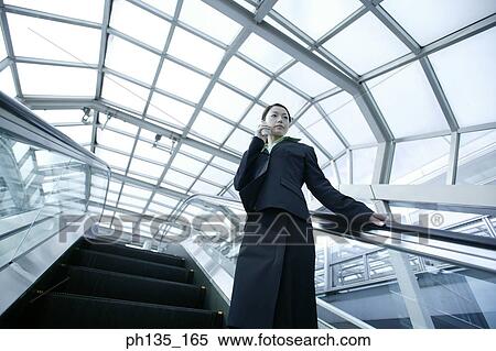 Stock Photography - Young businesswoman standing on an escalator talking on a mobile phone. Fotosearch