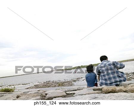 Rear view of a man and his son sitting near a river View Large Photo Image Picture - Rear view of a man and his son sitting near a river. Fotosearch