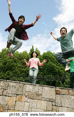 Low angle view of a young woman standing on a wall with two young men jumping View Large Photo Image Stock Photo - Low angle view of a young woman standing on a wall with two young men jumping. Fotosearch
