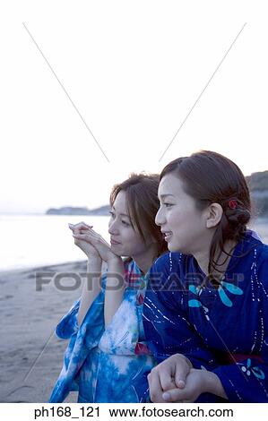 Stock Image - Two women sitting by the beach during sunset. Fotosearch