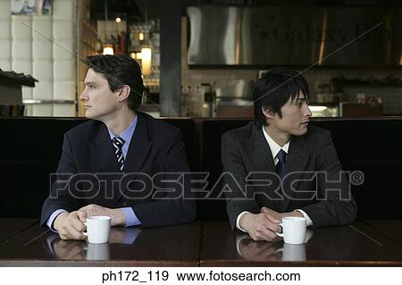 Two male executives sitting by table, holding a cup and looking in opposite direction View Large Photo Image Stock Photo - Two male executives sitting by table, holding a cup and looking in opposite direction. Fotosearch