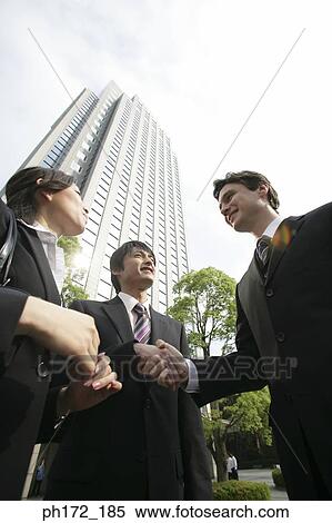 Two businessmen shaking hands with female executive looking on, low angle view View Large Photo Image Stock Photography - Two businessmen shaking hands with female executive looking on, low angle view. Fotosearch