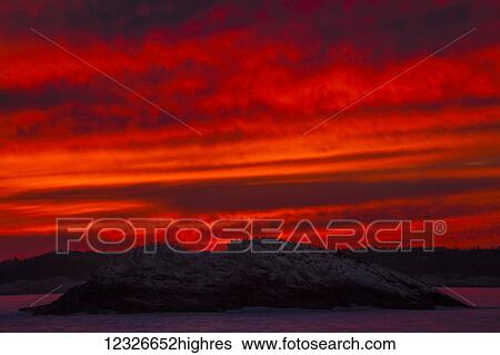 Stock Photograph - A blazing red sky at sunset silhouettes hundreds of Commorant seabirds on a rocky island as seen from the High Head Trail; Prospect, Nova Scotia, Canada. Fotosearch