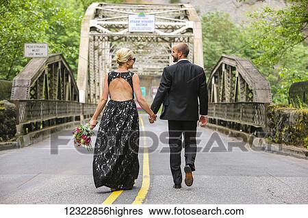 Stock Photograph - A bride and groom holding hands and walking down a paved road; Oregon, United States of America. Fotosearch