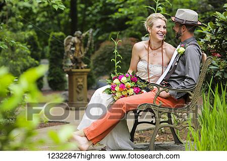 Stock Photograph - A bride and groom sit on a bench in a garden; Oregon, United States of America. Fotosearch