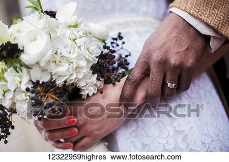 Stock Photograph - A bride and grooms hands together with a view of the wedding dress and flower bouquet; Portland, Oregon, United States of America. Fotosearch