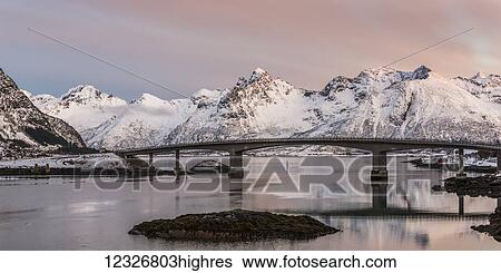 Stock Photograph - A bridge crossing the tranquil water with pink cloud over the rugged, snow covered mountains; Nordland, Norway. Fotosearch