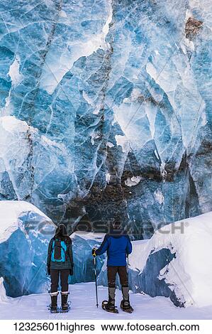 Stock Photograph - A couple of snowshoers examine a large blue wall of ice with several collapsed pieces of ice at the bottom at the terminus of Canwell Glacier in the Alaska Range; Alaska, United States of America. Fotosearch